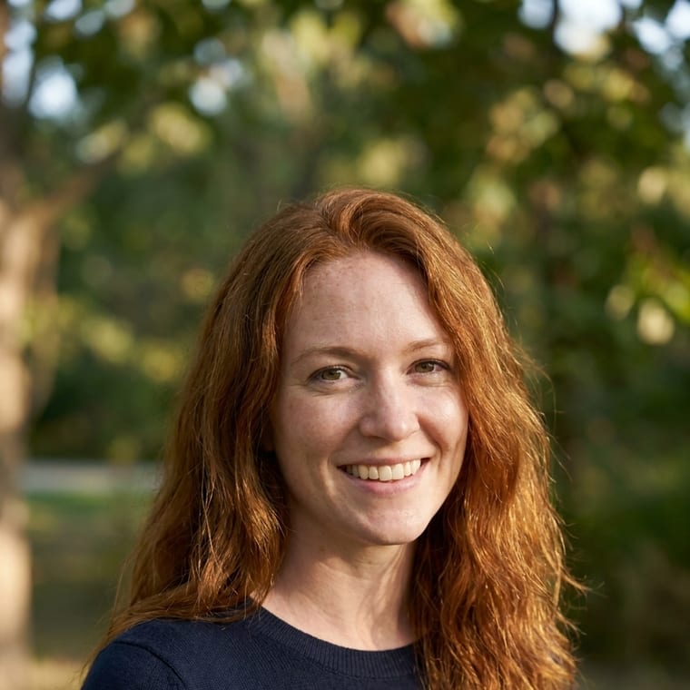 A person in a fine-gauge knit, outdoors with the background thrown fully out of focus, lit by soft window daylight, head tilted slightly toward the camera.