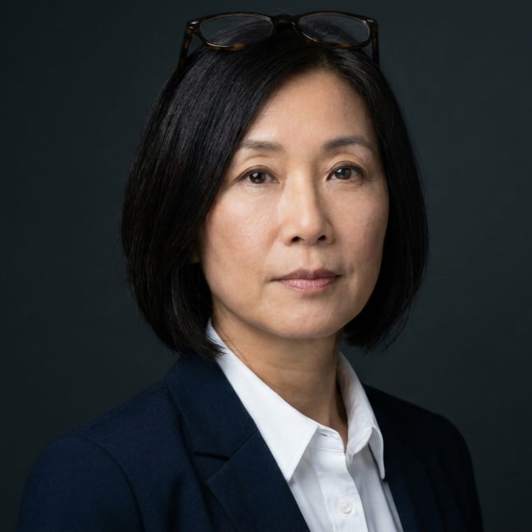 A woman in her early fifties in a dark blazer, on a charcoal seamless backdrop, lit with a Rembrandt key light.